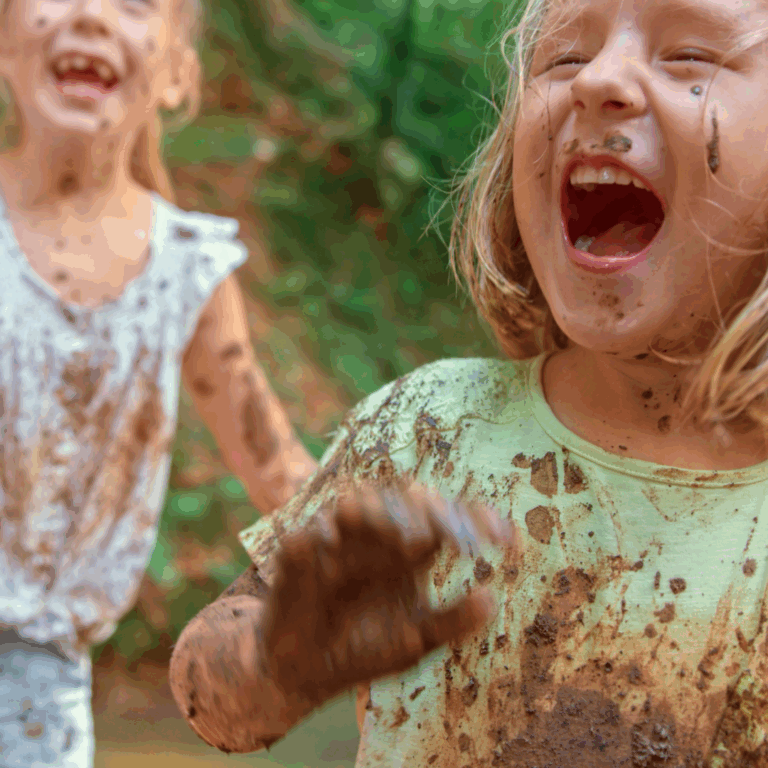 Preschoolers smiling with glee as they play in the mud kitchen with muddy faces and hands