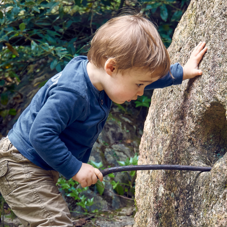 A child looking into a hole in a concrete object outdoors and using a stick to explore