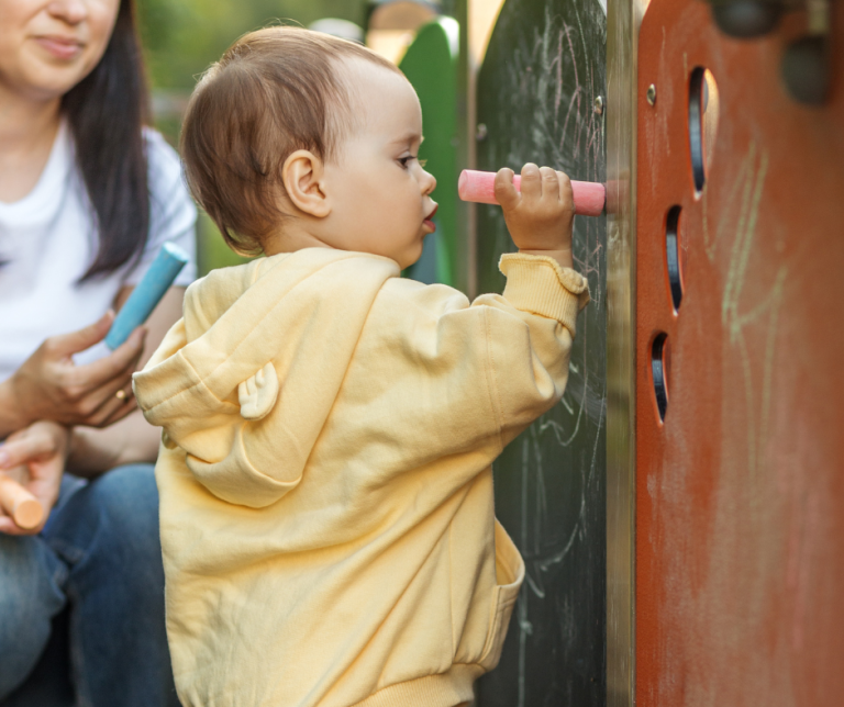 A child being supported by an adult to use chalk to mark make outdoors